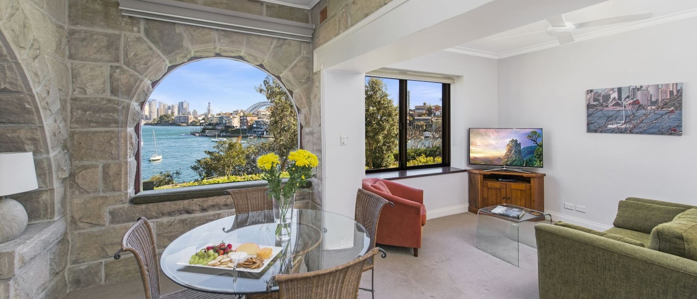 A living and dining space in a Sydney vacation rental, with stone walls in the dining area and a view from the window of Port Jackson and Sydney Harbour Bridge.