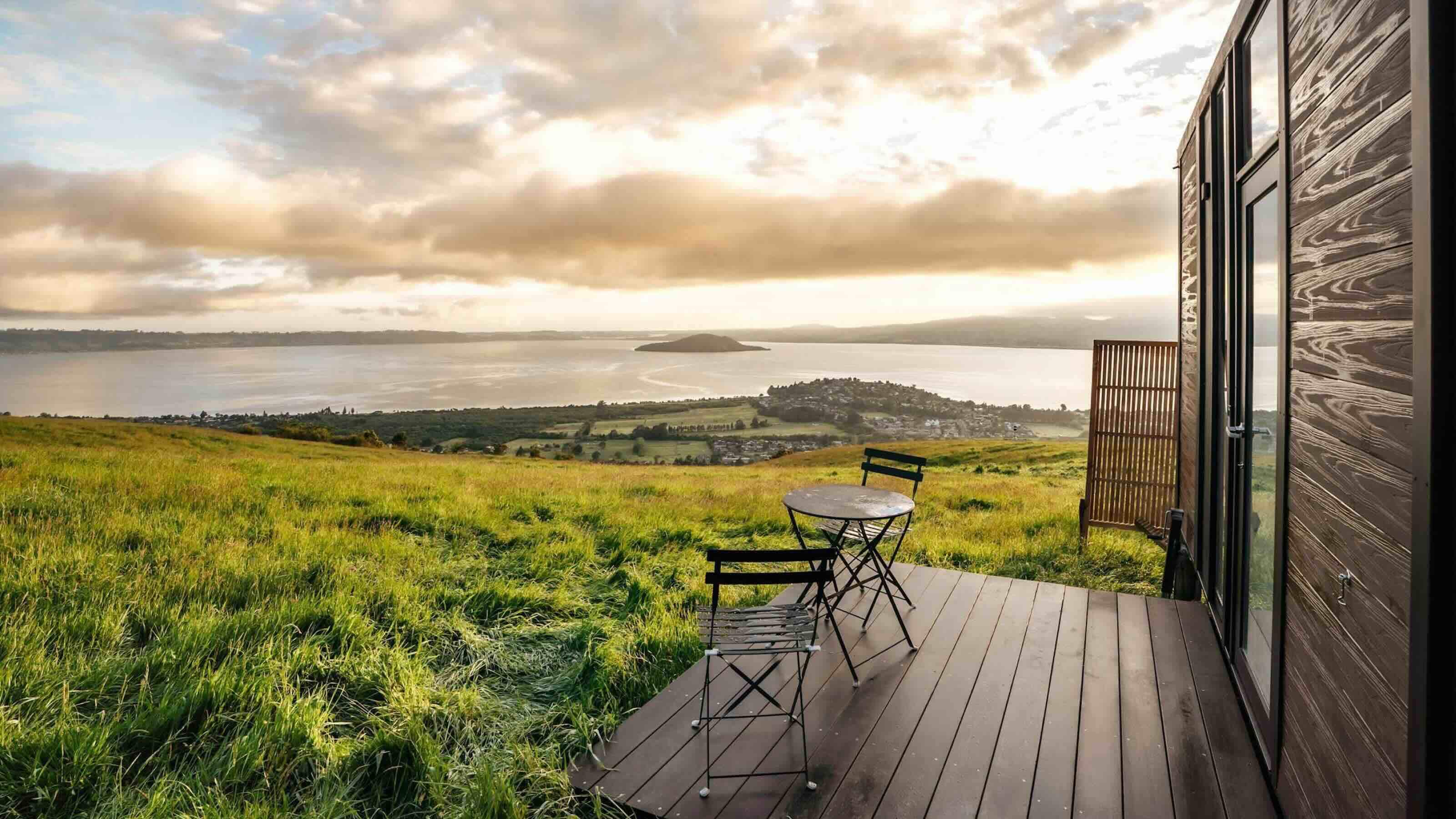 Two dark wood chairs and a table sit on the porch of a wooden cabin with a view of the Bay of Plenty in New Zealand’s North Island.  