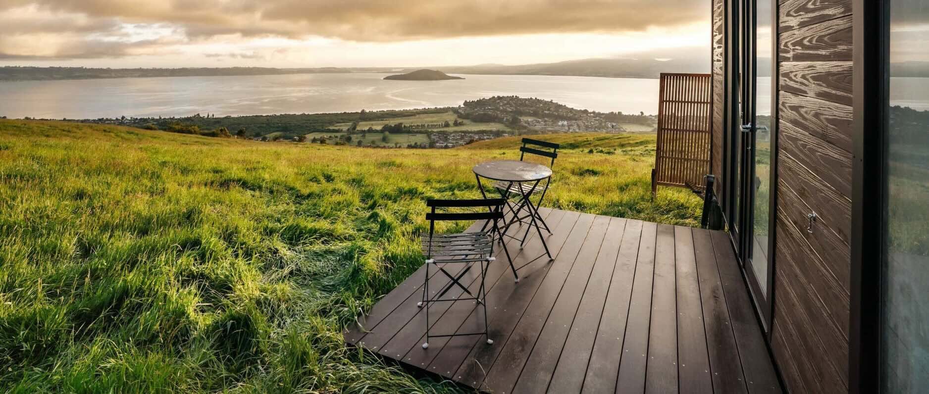Two dark wood chairs and a table sit on the porch of a wooden cabin with a view of the Bay of Plenty in New Zealand’s North Island. 