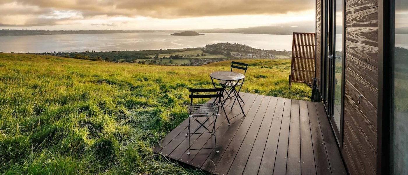 Two dark wood chairs and a table sit on the porch of a wooden cabin with a view of the Bay of Plenty in New Zealand’s North Island.