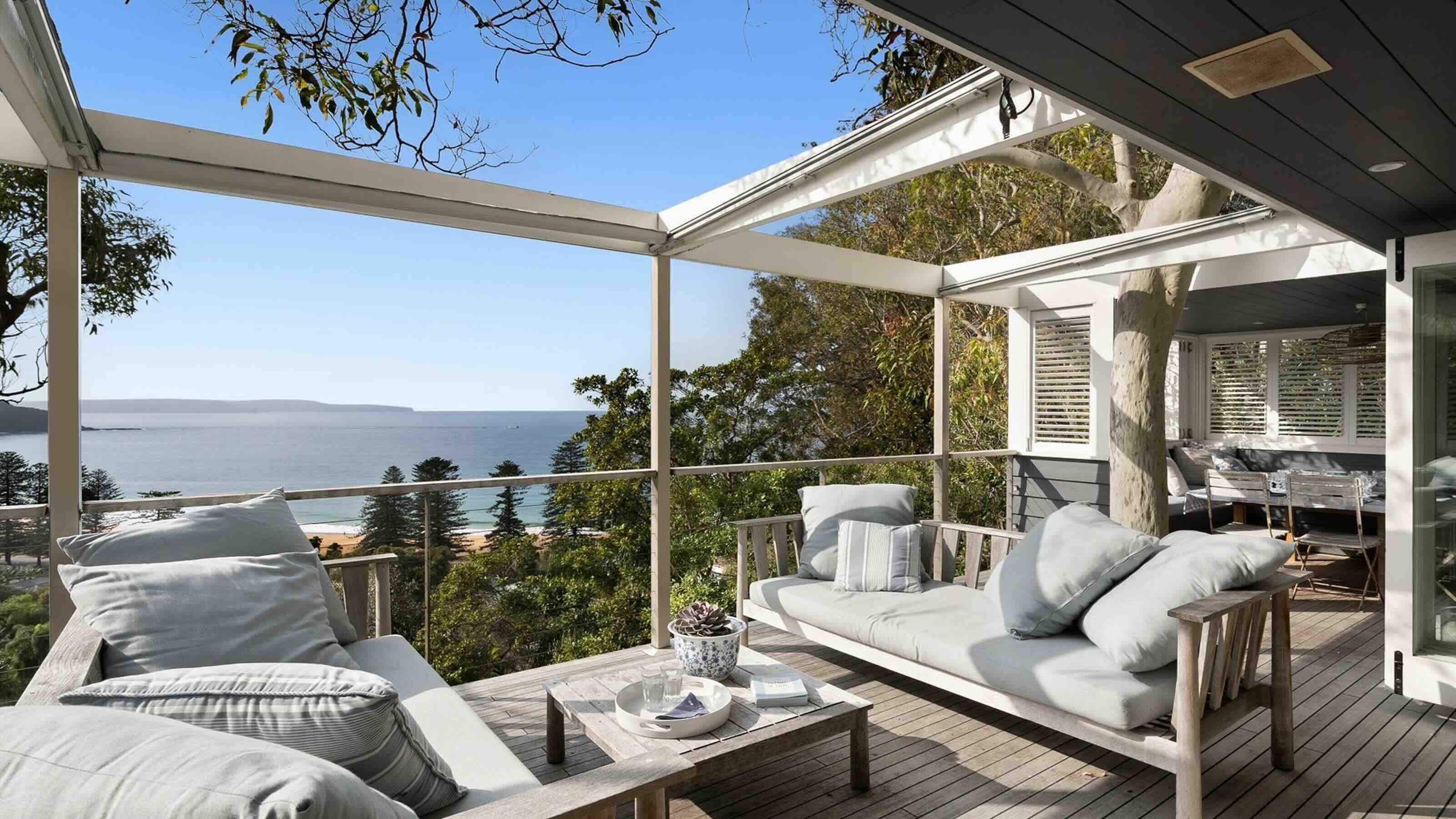 The spacious wooden deck of a holiday home in Palm Beach, Australia. Two outdoor sofas are positioned opposite each other with a table in the middle. A covered outdoor dining area is visible behind one sofa. 