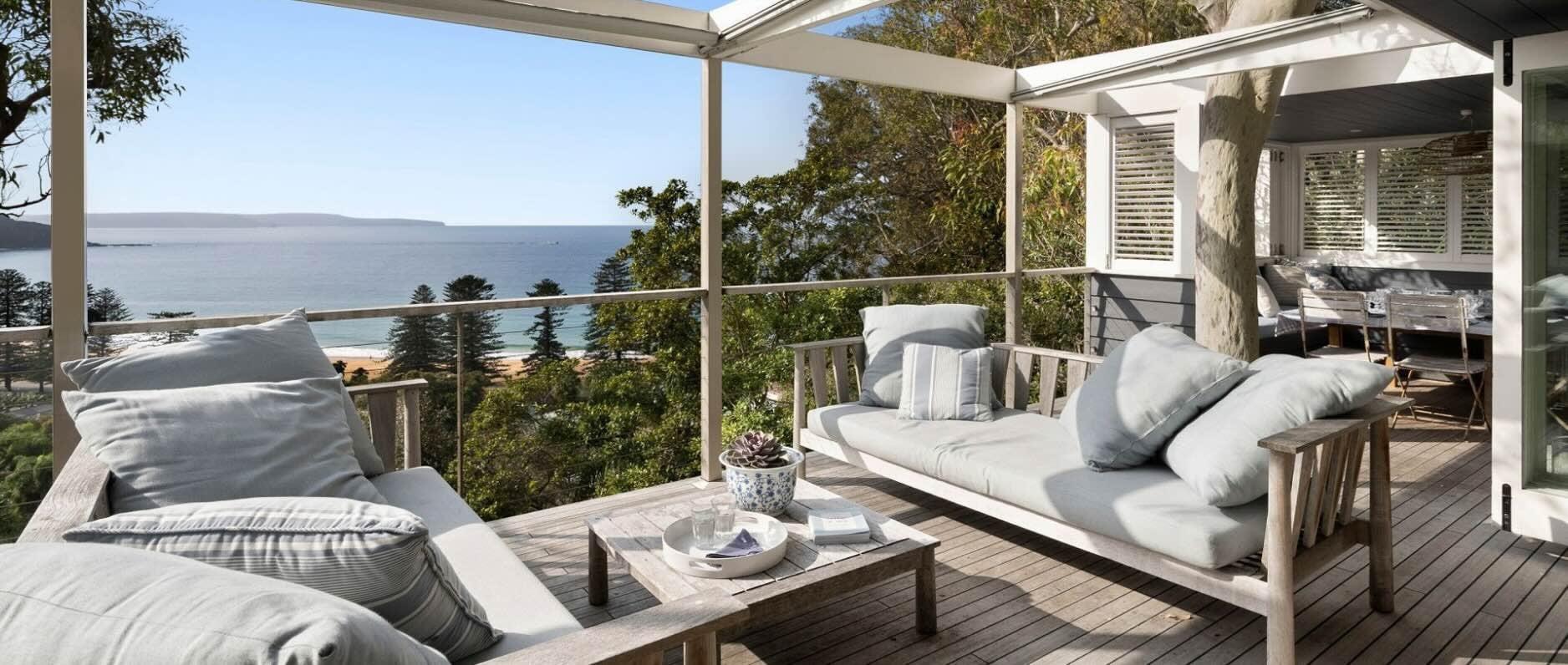 The spacious wooden deck of a holiday home in Palm Beach, Australia. Two outdoor sofas are positioned opposite each other with a table in the middle. A covered outdoor dining area is visible behind one sofa. 