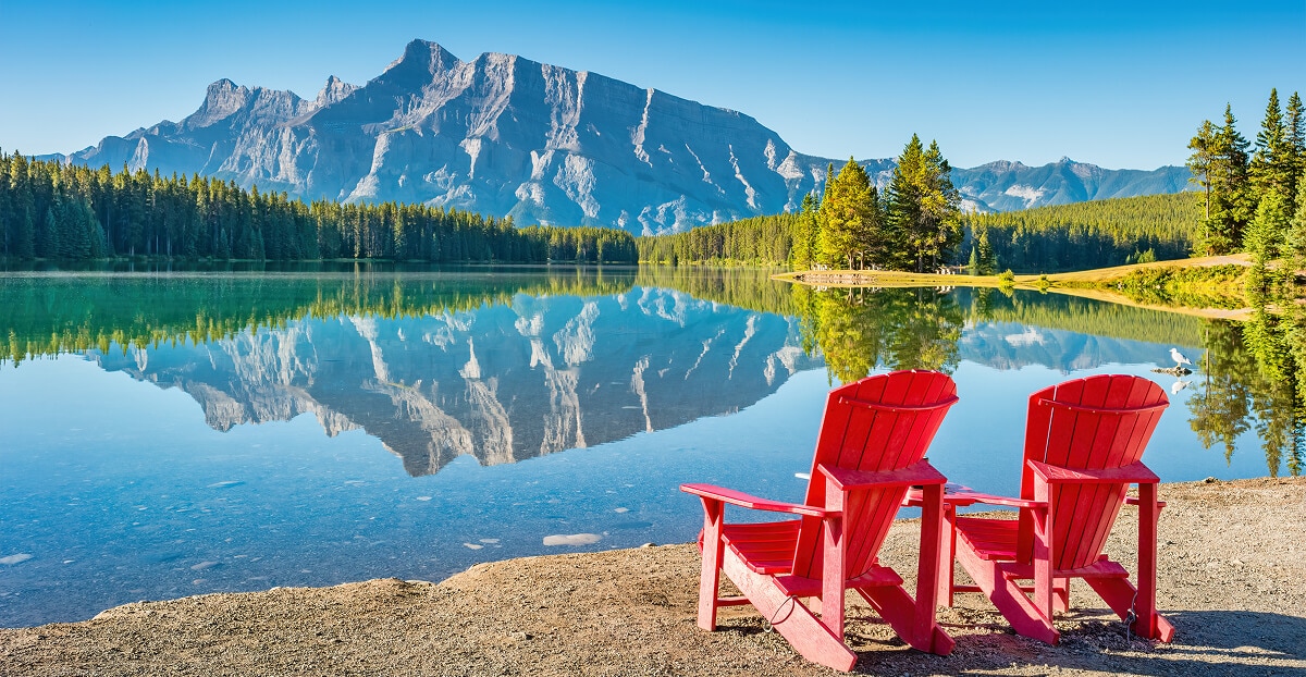 Two red Muskoka chairs are positioned on the natural shoreline of Two Jack Lake in Banff National Park. Mount Rundle rises from the skyline and is reflected in the lake’s tranquil waters.