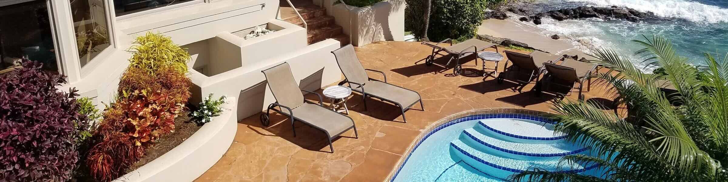 An aerial view of a luxury oceanfront villa with solar panels on the roof, a stone patio featuring a swimming pool and lounge chairs, and waves crashing against the rocky shoreline.