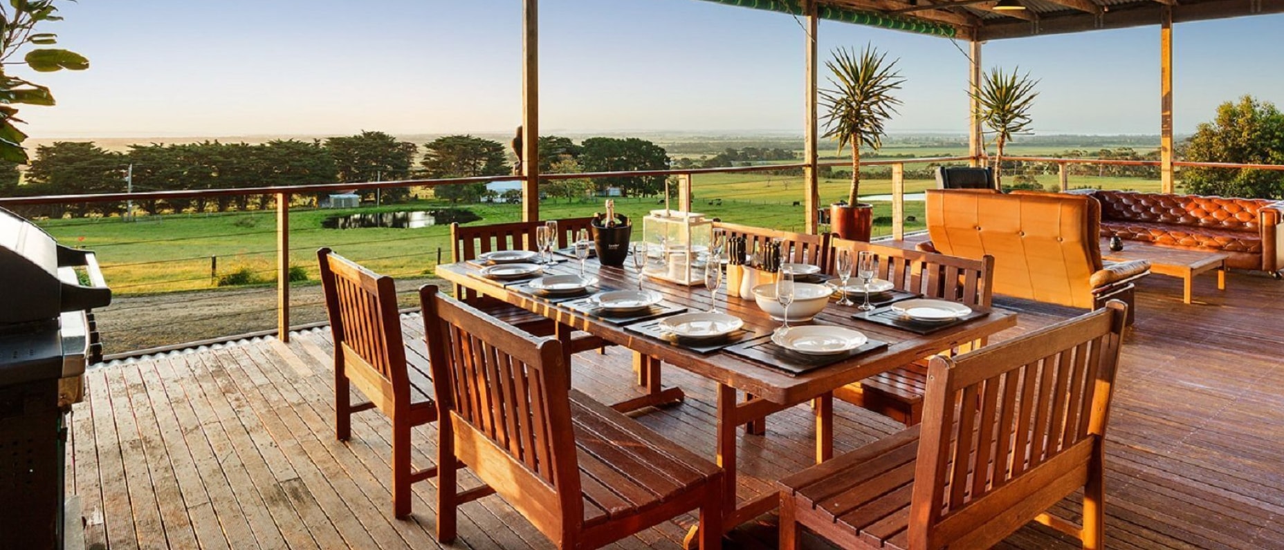 A spacious, covered wooden deck overlooks tranquil green fields and lakes at sunset. A large wooden dining table in the foreground is set for an evening meal, with a barbecue behind. In the background, a cosy outdoor lounge area features a two-seater couch, armchair and Chesterfield sofa. 