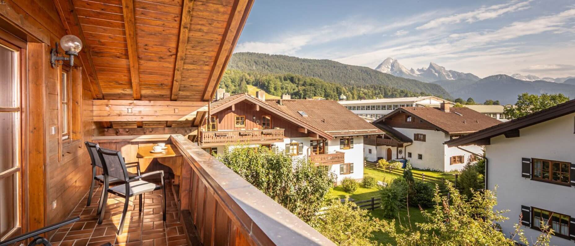 Ein sonniger Holzbalkon eines rustikalen Chalets blickt auf ein Tal mit traditionellen Häusern, dunklen Dächern und grünen Gärten. Im Hintergrund erheben sich Alpengipfel hinter dicht bewaldeten Hügeln. Der Balkon hat Terrakottafliesen, drei schwarze Netzstühle und einen kleinen Tisch mit zwei Teetassen und Untertassen.