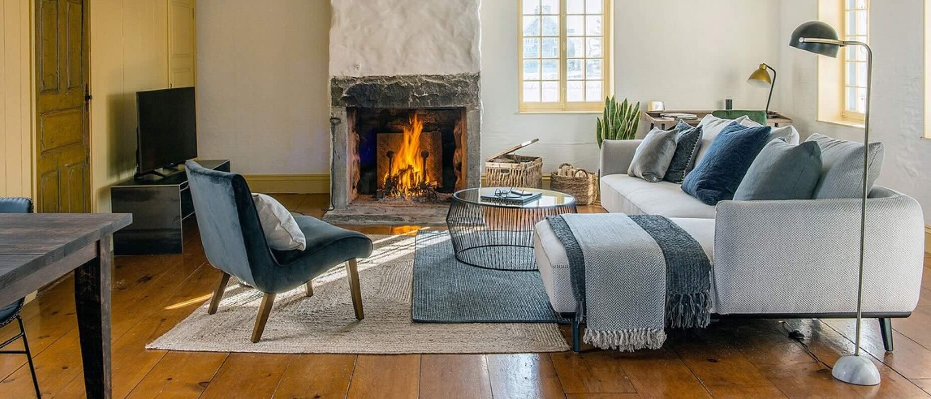 Un salon rustique doté d’un grand foyer en pierre, où crépite un feu. Un sofa modulaire gris clair se trouve à droite de la pièce, orné de couvertures et de coussins bleus et gris. Un fauteuil d’appoint en velours est placé face au foyer, et une table métallique noire trône au centre de la pièce.