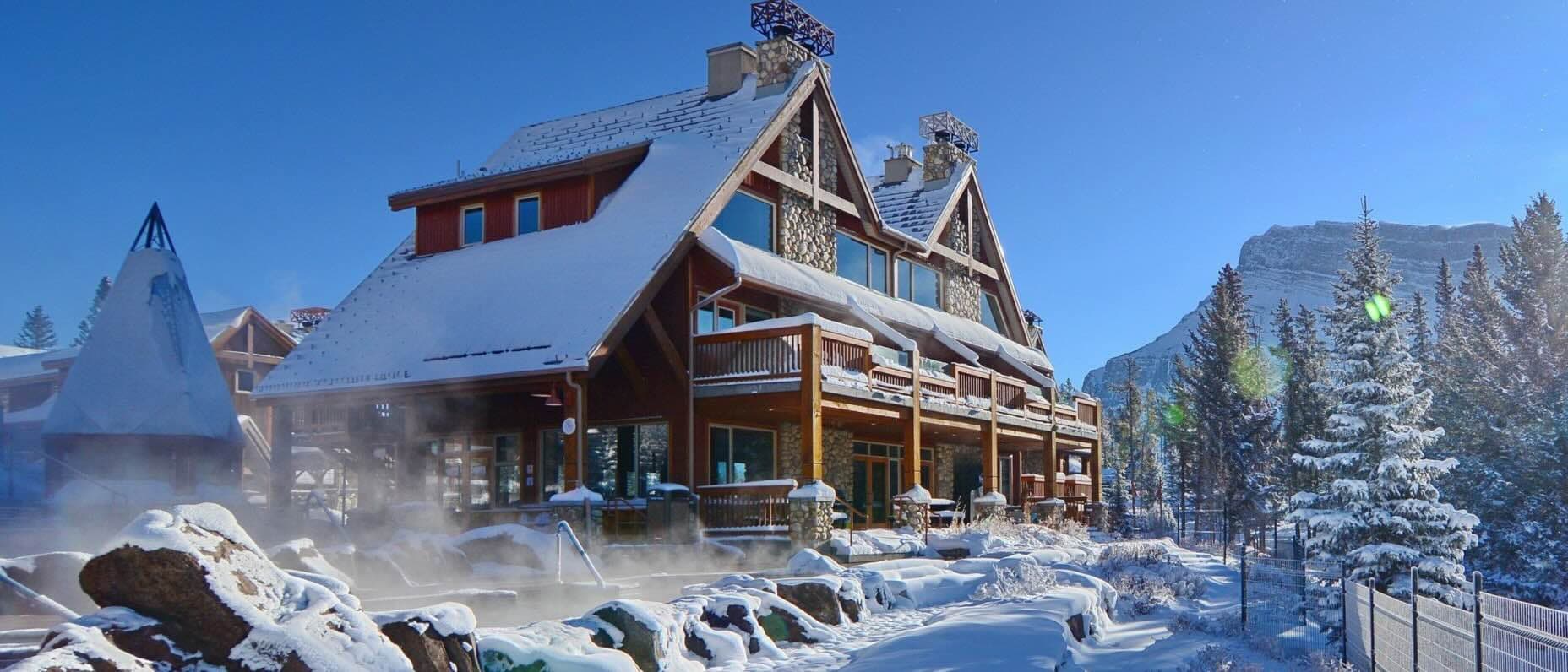 Location de vacances haut de gamme dans un chalet de montagne en hiver, avec des sources thermales fumantes à l’avant, une architecture en pierre et en bois, et les sommets spectaculaires des Rocheuses en arrière-plan.