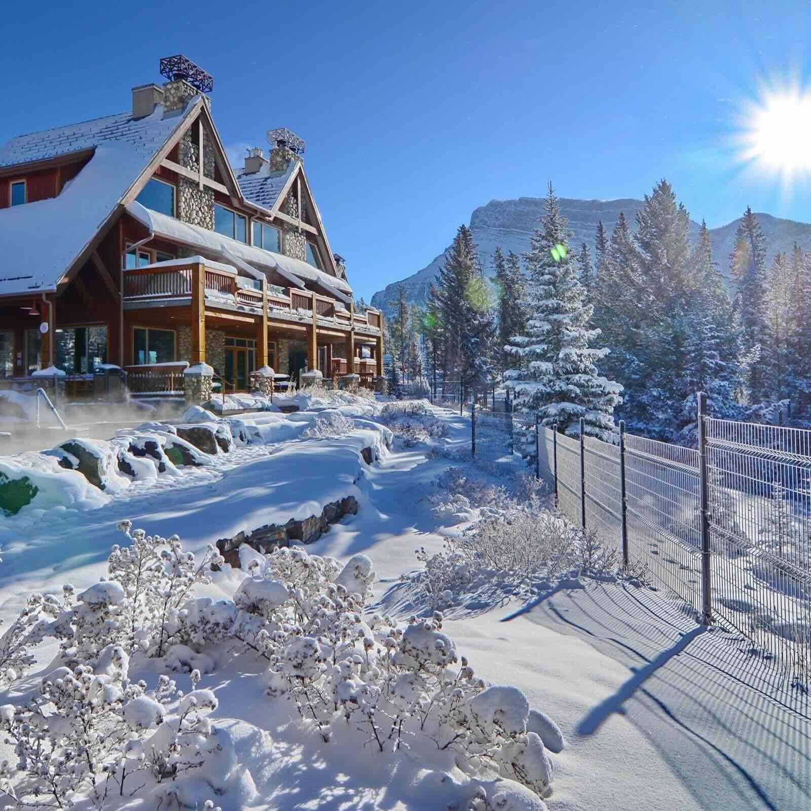 Location de vacances haut de gamme dans un chalet de montagne en hiver, avec des sources thermales fumantes à l’avant, une architecture en pierre et en bois, et les sommets spectaculaires des Rocheuses en arrière-plan.