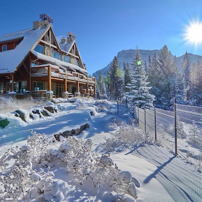 Location de vacances haut de gamme dans un chalet de montagne en hiver, avec des sources thermales fumantes à l’avant, une architecture en pierre et en bois, et les sommets spectaculaires des Rocheuses en arrière-plan.