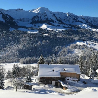 Gemütliches alpines Chalet als Ferienunterkunft, eingebettet in ein verschneites Tal mit weitem Blick auf die Berge und mit Raureif überzogenen Kiefern an einem klaren Wintertag