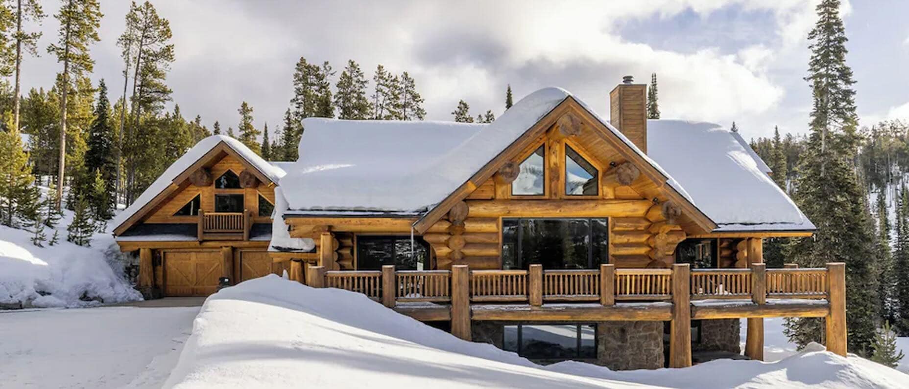 Luxury log cabin vacation rental covered in snow with a wraparound deck, floor-to-ceiling windows, and pine forest backdrop — perfect for a mountain winter getaway