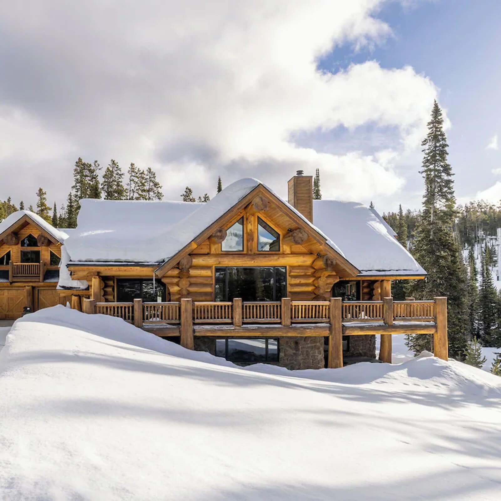Luxury log cabin vacation rental covered in snow with a wraparound deck, floor-to-ceiling windows, and pine forest backdrop — perfect for a mountain winter getaway