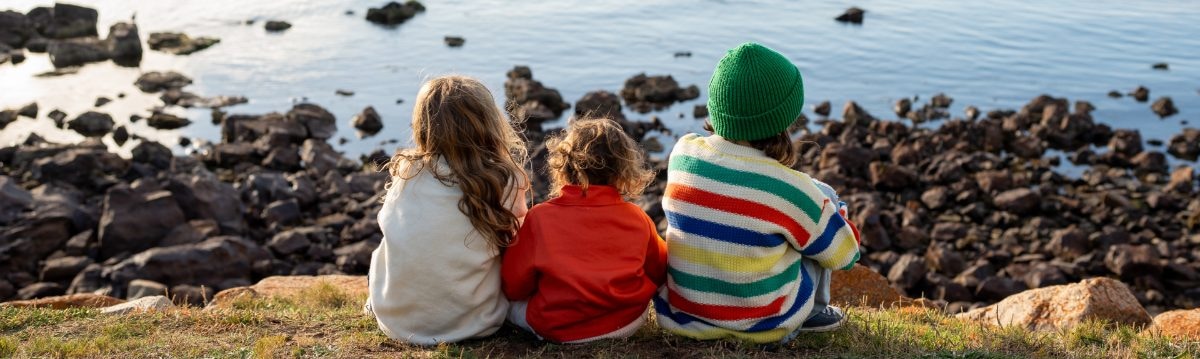 Three young children dressed in cozy fall outfits sit side-by-side on a grassy bank. They look out over a rocky coastline and towards the sea.
