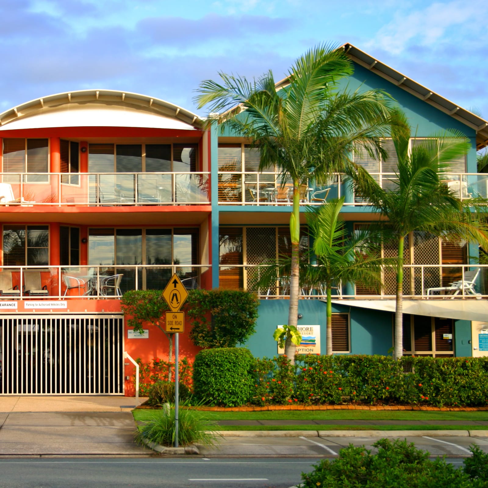 A front-facing view from the road of three-storey teal and orange tropical resort homes. The homes feature glass-fronted balconies, filled with seats and sun loungers. Lush palm trees and greenery sit in front of the houses.
