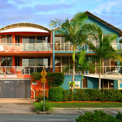 A front-facing view from the road of three-storey teal and orange tropical resort homes. The homes feature glass-fronted balconies, filled with seats and sun loungers. Lush palm trees and greenery sit in front of the houses.