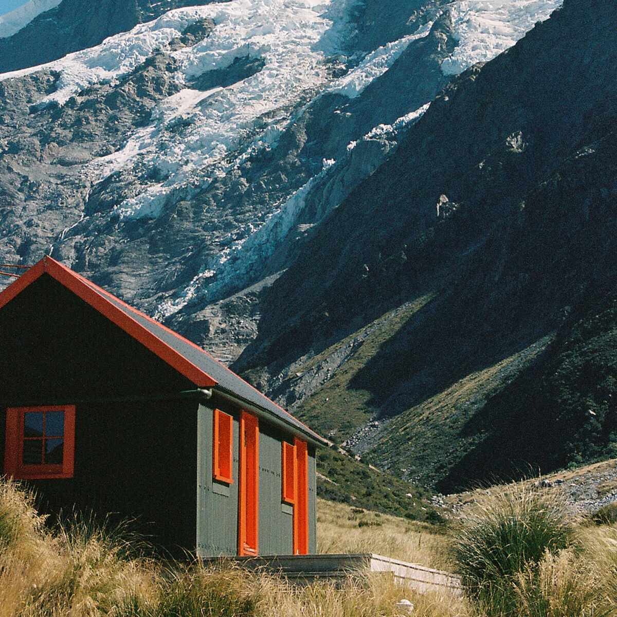 A dark green wooden mountain cabin with a bright red roof trim, window shutters, and door frame stands in a grassy alpine valley. A small wooden deck wraps around the front of the cabin. In the background, a snow-covered mountain rises dramatically.