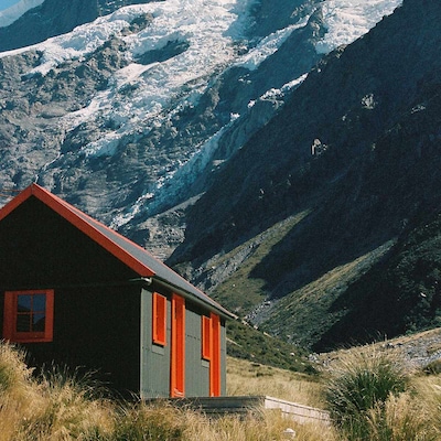A dark green wooden mountain cabin with a bright red roof trim, window shutters, and door frame stands in a grassy alpine valley. A small wooden deck wraps around the front of the cabin. In the background, a snow-covered mountain rises dramatically.