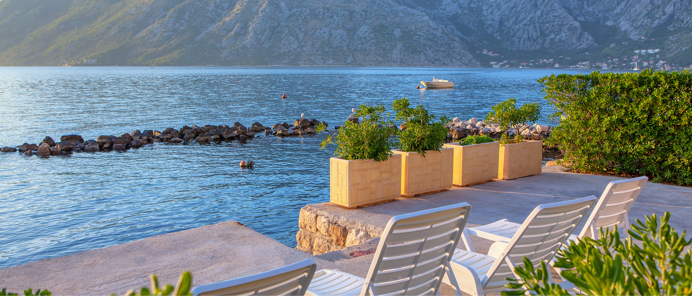 Four white lounge chairs are positioned on a stone terrace with four plant pots with shrubs in front. The terrace looks over an open stretch of water with mountains rising in the background.
