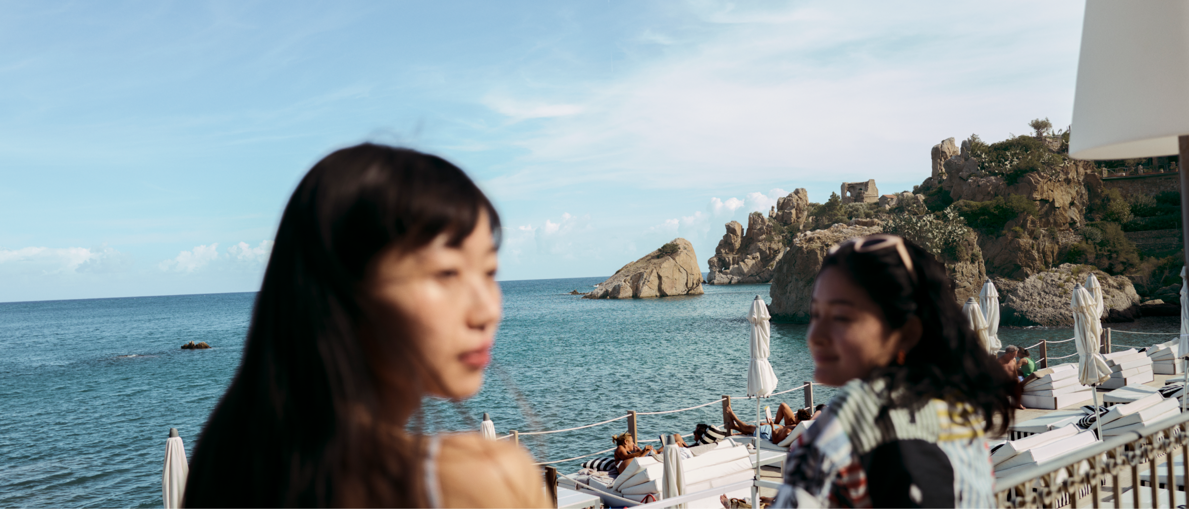 Two women stand atop a terrace overlooking a busy pool area. Beyond the pool area is a calm sea and distant rock formations.