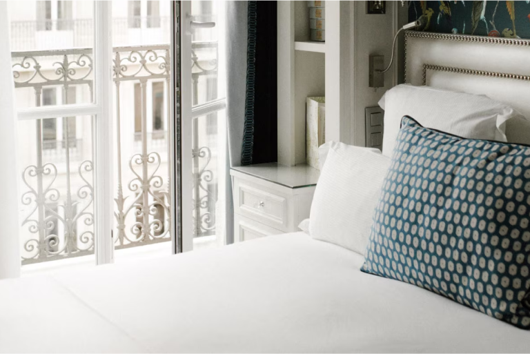 A hotel interior reveals a bed with white linen and a blue and white polka dot pillow. French doors leading to a balcony are slightly ajar.