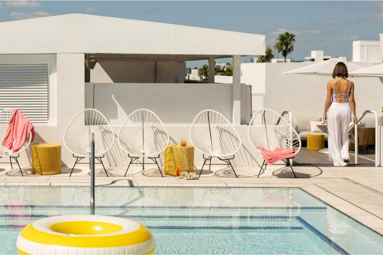 Patio chairs line a swimming pool with a citrus yellow inner tube floating in it. A woman in a spaghetti strap swim top and white linen pants exits the pool area.