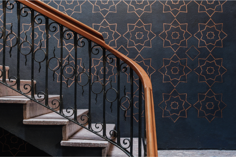 An upclose shot of a marble staircase reveals a woodern bannister and ornate ironwork. The background wallpaper has a stencil pattern.