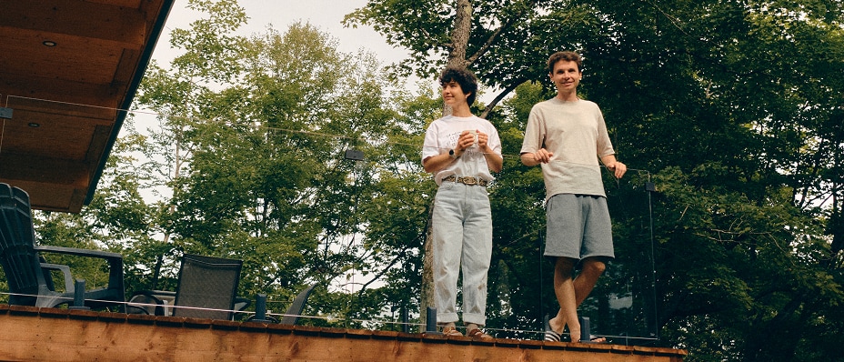 Two people stand on a raised wooden deck beside a modern house, surrounded by tall trees.