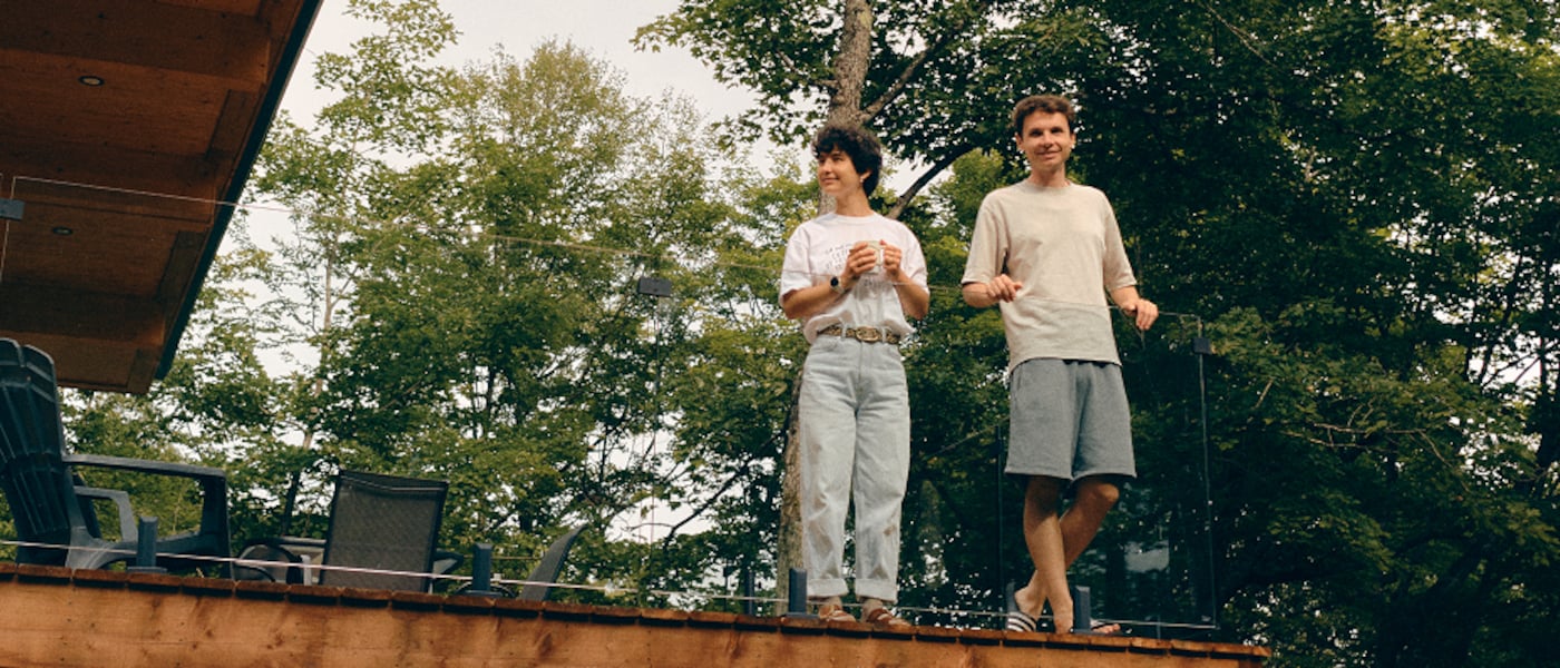 Two people stand on a raised wooden deck beside a modern house, surrounded by tall trees.