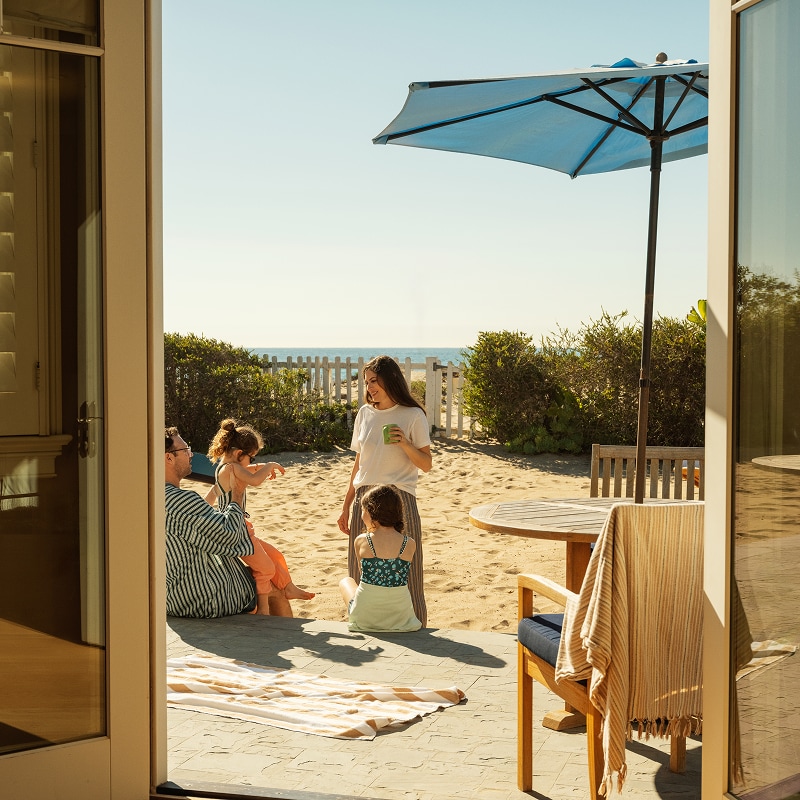 A family relaxing on a sandy patio bordered by a white picket fence that leads directly onto a beach