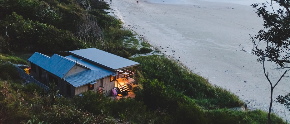 A cozy beach house glows at dusk beside a quiet sandy shoreline surrounded by lush greenery.