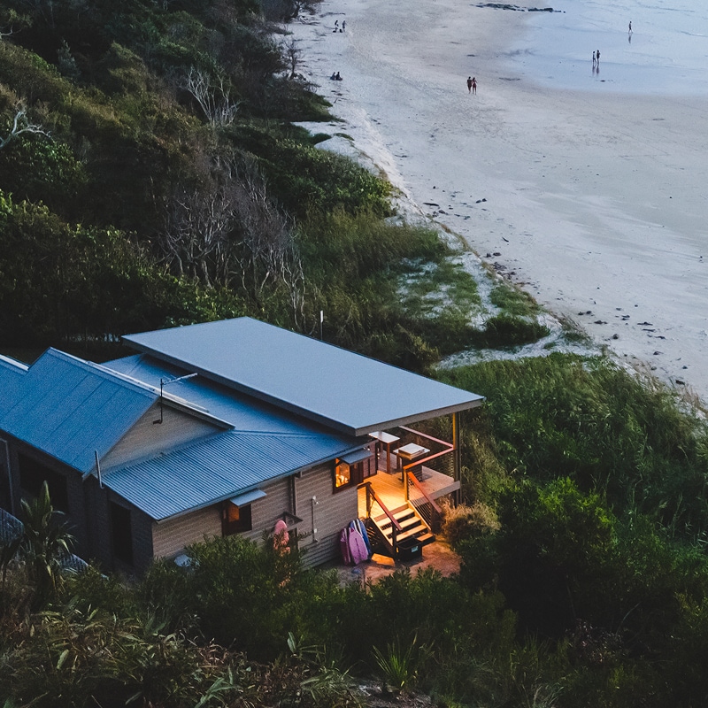 A cozy beach house glows at dusk beside a quiet sandy shoreline surrounded by lush greenery.