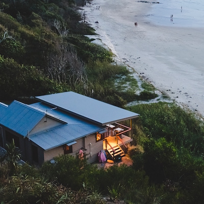 A cozy beach house glows at dusk beside a quiet sandy shoreline surrounded by lush greenery.