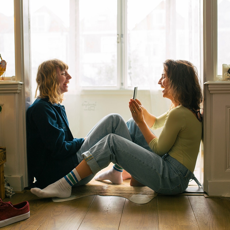 Deux femmes assises près d’une fenêtre ensoleillée sourient en partageant un moment de détente à l’intérieur.