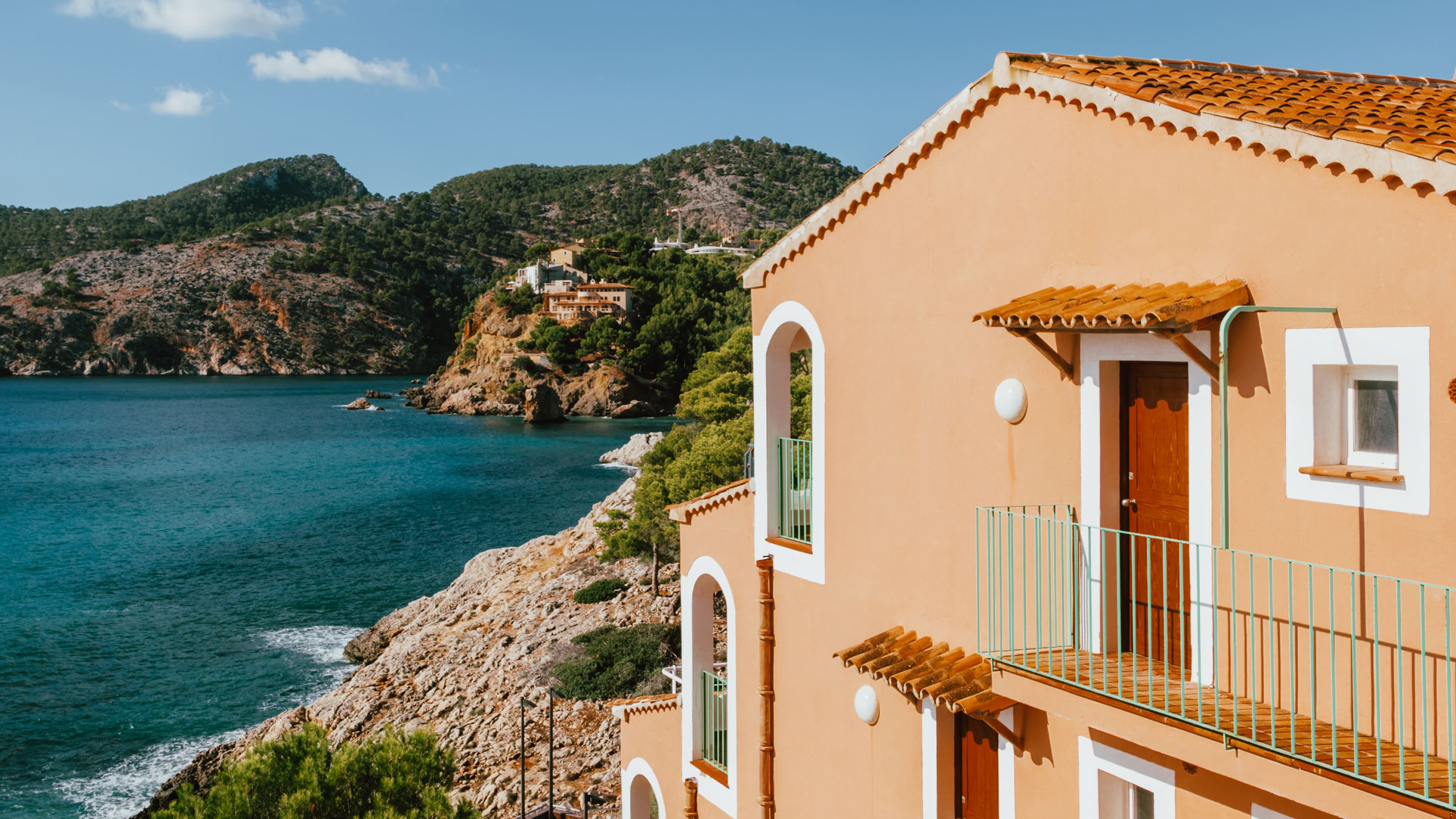 Eine sonnenbeschienene Villa am Meer blickt auf klares blaues Wasser und raue Küstenhügel.