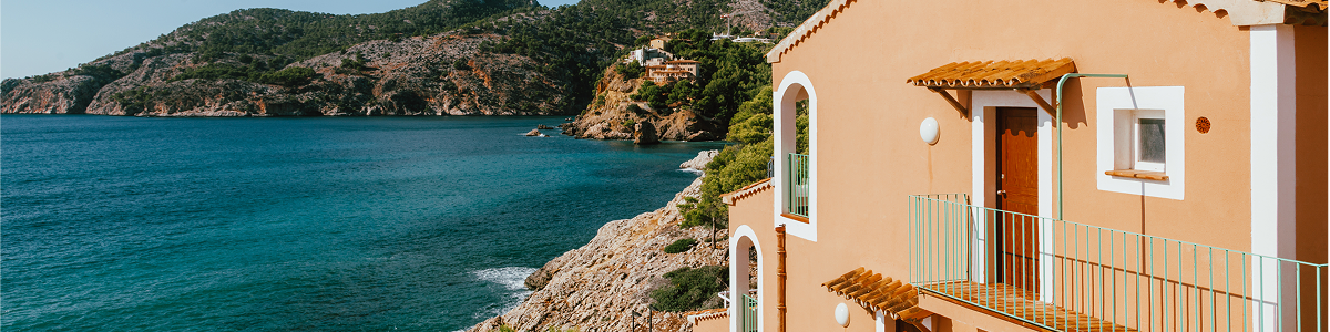 Eine sonnenbeschienene Villa am Meer blickt auf klares blaues Wasser und raue Küstenhügel.