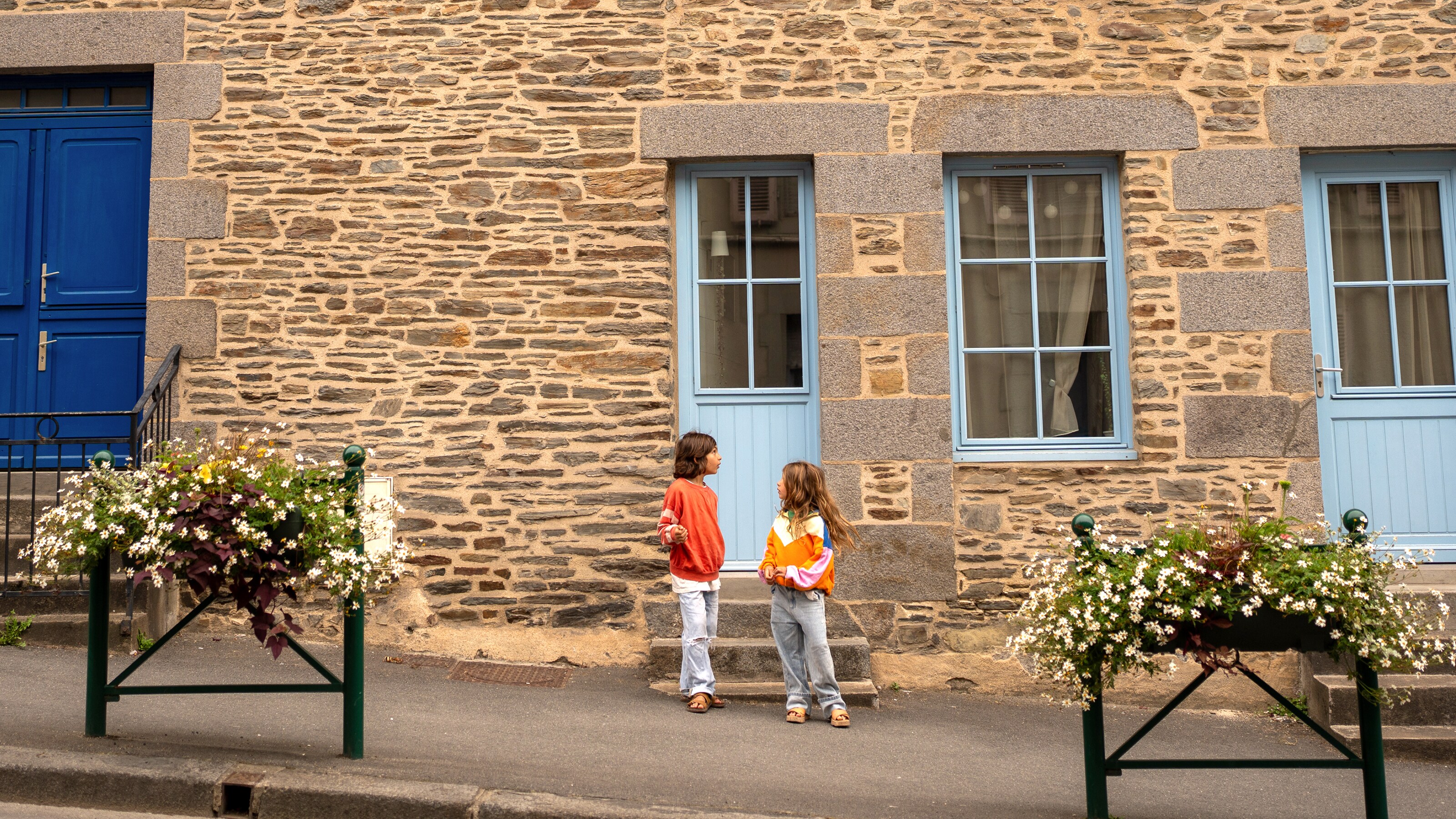 Deux enfants discutent dans une rue calme devant une maison en pierre rustique aux portes et fenêtres bleu vif.