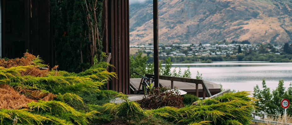 A modern timber cottage with a deck overlooks a tranquil lake and mountains, surrounded by lush greenery.