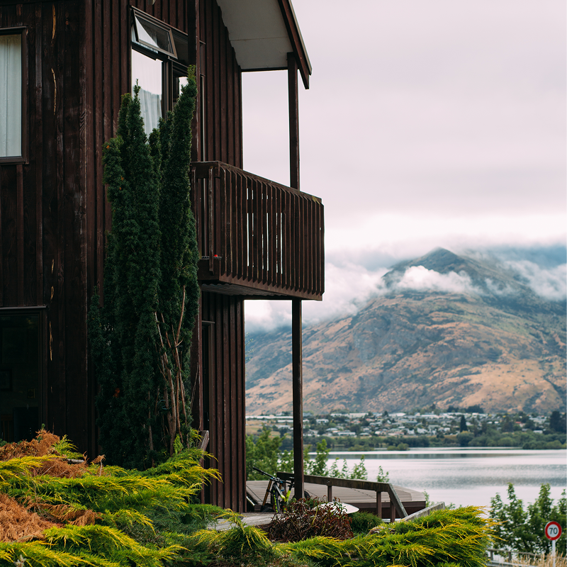 A modern timber cottage with a deck overlooks a tranquil lake and mountains, surrounded by lush greenery.