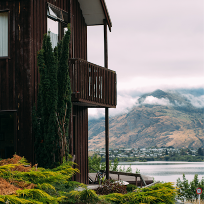 A modern timber cottage with a deck overlooks a tranquil lake and mountains, surrounded by lush greenery.