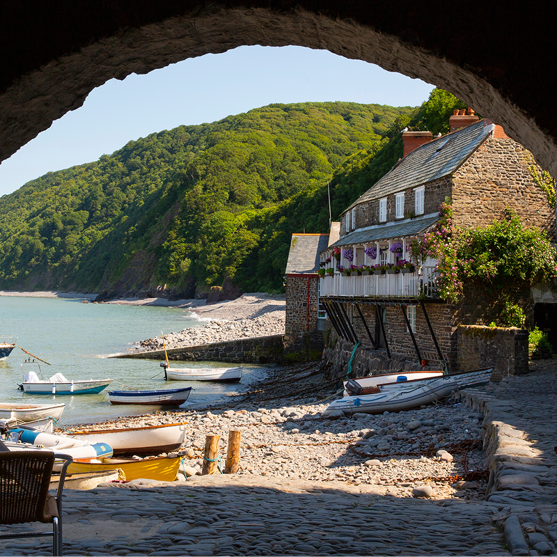 A stone cottage and small boats sit along a peaceful cove framed by a rustic archway and green hills.