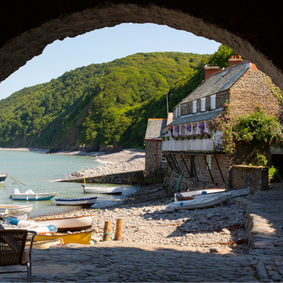 A stone cottage and small boats sit along a peaceful cove framed by a rustic archway and green hills.