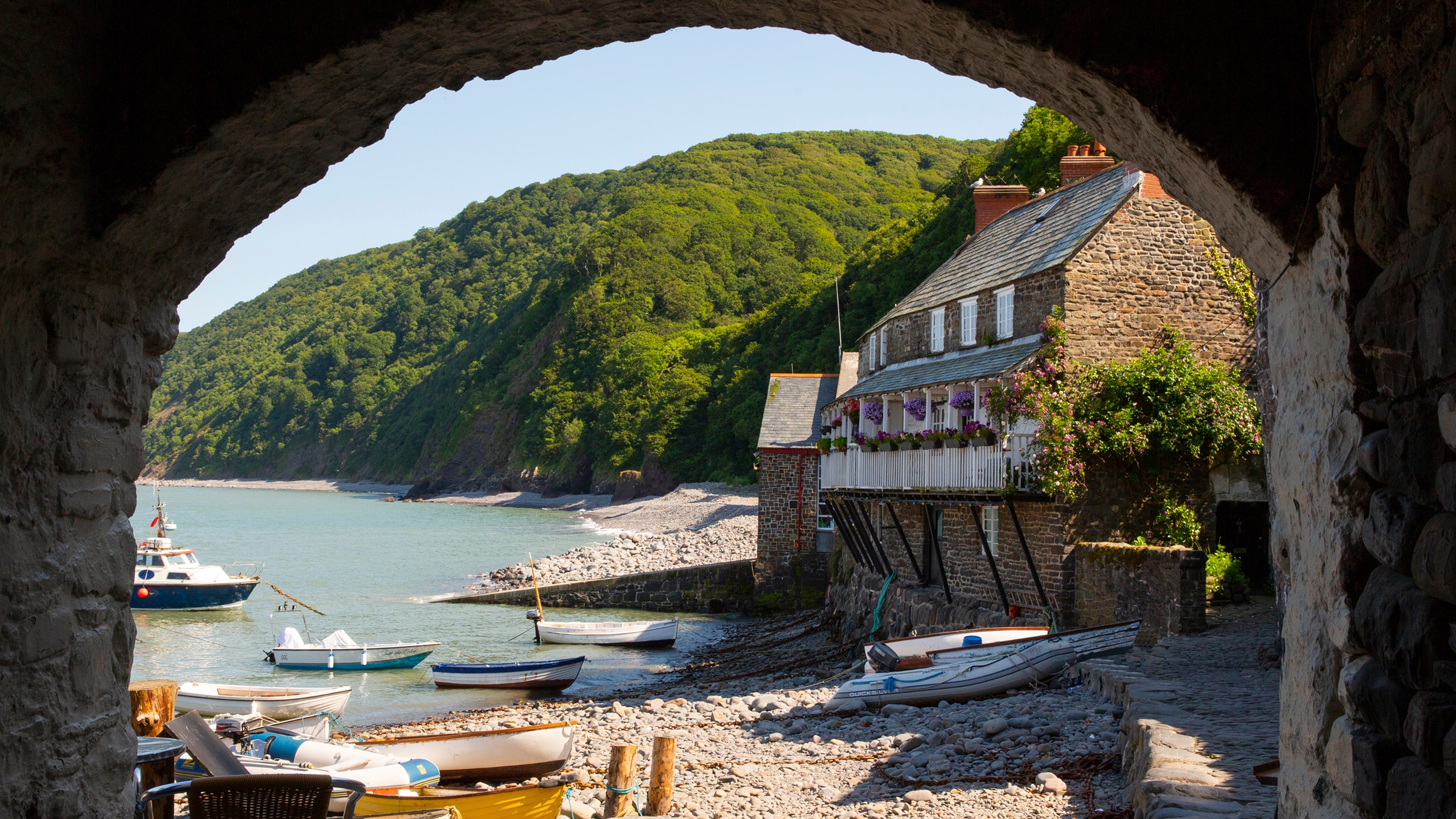A stone cottage and small boats sit along a peaceful cove framed by a rustic archway and green hills.