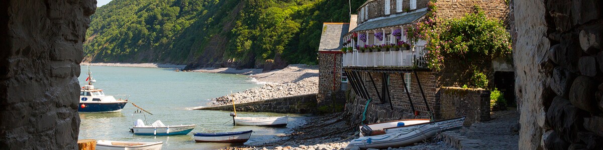 A stone cottage and small boats sit along a peaceful cove framed by a rustic archway and green hills.