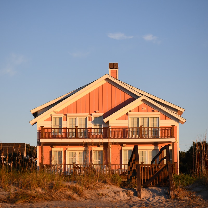 An inviting beach house glows in warm sunlight behind sandy dunes and sea grass.