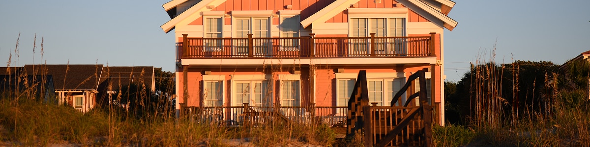 An inviting beach house glows in warm sunlight behind sandy dunes and sea grass.