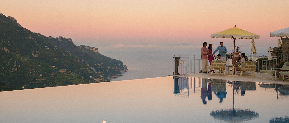 En haut d’une falaise surplombant la côte amalfitaine, une piscine à débordement reflète un ciel aux teintes pastel au coucher du soleil, se fondant harmonieusement dans la vue sur l’océan en contrebas. À droite de la piscine, les voyageurs sont rassemblés autour de chaises de patio jaunes, disposées sous un parasol jaune.