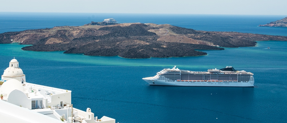 A large white cruise ship sails through the turquoise waters near Santorini, Greece, with a volcanic island in the background and classic whitewashed buildings in the foreground.