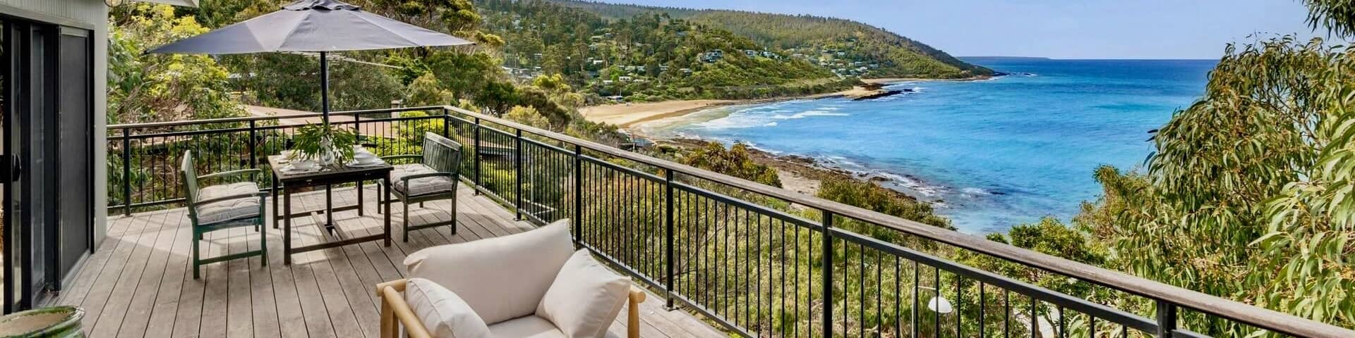 A wooden deck with chairs and a table with a grey umbrella faces a tree-covered beach and an aqua ocean.