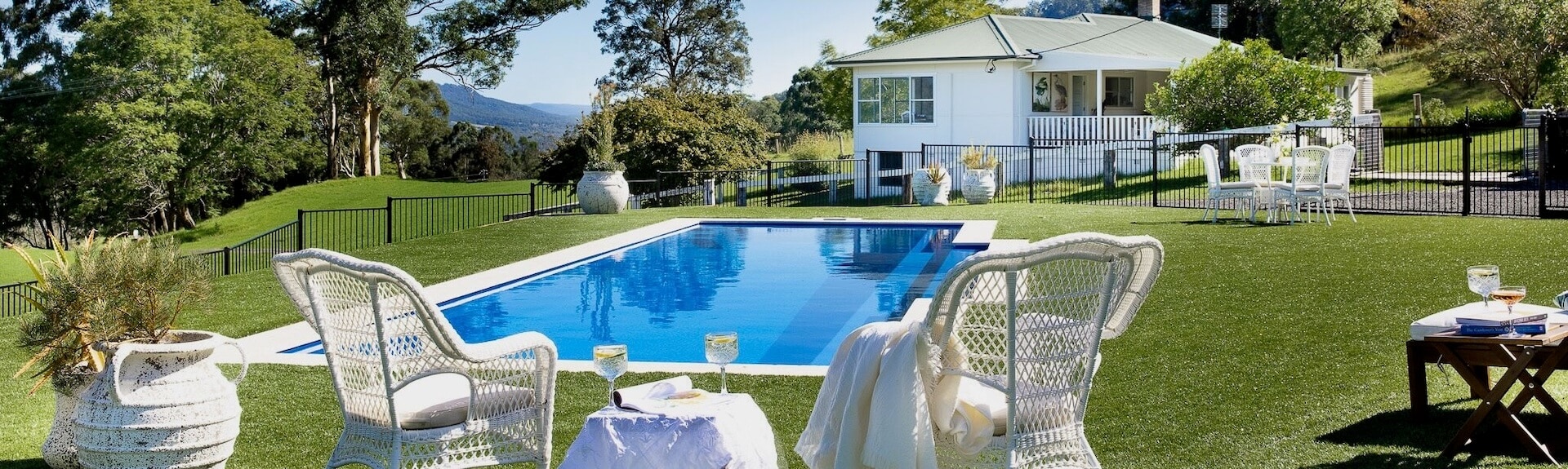 A private holiday home with a green lawn and an outdoor pool surrounded by scattered seating and a black bar fence. There is blue sky and mountains in the distance. 